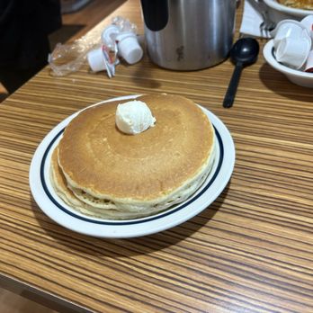 IHOP - A white plate holds three stacked pancakes topped with a small dollop of butter. The pancakes are golden brown and appear fluffy. The setting is a restaurant with a wooden table, and in the background, there are small containers of cream or syrup, a metal pitcher, and a bowl with more cream packets. - Breakfast & Brunch Near Me - Union, New Jersey