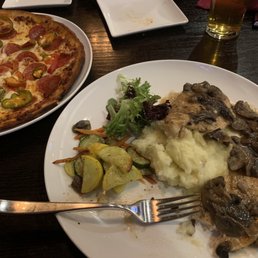 Chicken Marsala and pizza - A dinner plate contains chicken topped with mushrooms, served with mashed potatoes and a side of sautéed vegetables including zucchini and carrots. A small salad with mixed greens is also on the plate. Next to the plate, there is a pizza with pepperoni, green peppers, and other toppings. A fork rests on the edge of the plate, and a glass of beer is visible in the background. The setting appears to be a casual restaurant with a dark wooden table.