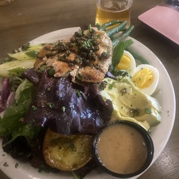 Circle D Eatery - Trout Nicoise Salad - A plate of mixed greens salad includes grilled chicken topped with herbs, boiled eggs sliced in half, roasted potato, and various leafy greens. A small container of dressing is placed on the side. A glass of beer is positioned behind the plate on a wooden table, with a pink item and other objects partially visible. - Coffee & Tea Near Me - Escalante, Utah