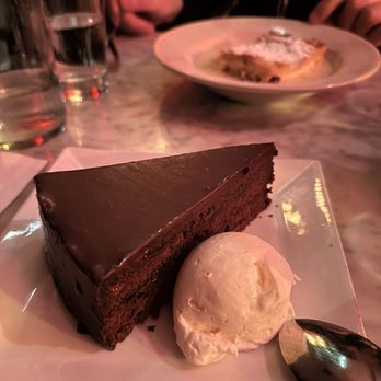 Werkstatt - Sacher Torte - A slice of chocolate cake with a smooth, glossy chocolate glaze is presented on a white square plate. Next to the cake, there is a scoop of vanilla ice cream. The setting appears to be a restaurant with a marble table, and in the background, there are glasses of water and a person with a plate of food. The lighting is warm and dim. - Austrian Near Me - Brooklyn, New York