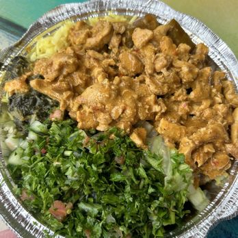 Marhaba - The image shows a round, foil takeout container filled with a serving of chicken curry topped with a yellowish sauce, accompanied by chopped greens and shredded lettuce. The container is placed on a multicolored surface, suggesting a casual dining or takeout setting. The food appears freshly prepared, with the greens and lettuce providing fresh side components to the main dish. - Mediterranean Near Me - McLean, Virginia