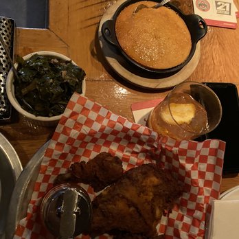 The Eagle OTR - 1/2 Chicken + greens + cornbread - The image shows a meal served on a wooden table, including fried chicken pieces on a checkered paper-lined tray, a bowl of cooked greens, a small skillet of gravy or sauce, and a glass of iced beverage with lemon. The setting appears to be a casual dining restaurant with a rustic atmosphere, and there are some business cards or promotional materials visible on the table. - American Near Me - Cincinnati, Ohio