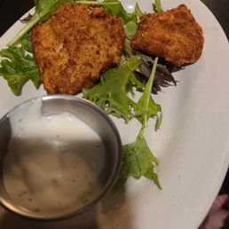bison balls - The image shows a white oval plate with two breaded and fried food items, accompanied by fresh mixed greens. A small metal cup containing a creamy dipping sauce is placed on the plate. The setting appears to be a restaurant, with a dark table surface and a glass dish visible in the background. The presentation suggests a dish served with a side of greens and a dipping sauce, typical of upscale or casual dining establishments.