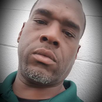 The Joint Chiropractic - A close-up photo of a man with a serious expression, short hair, and a goatee, standing against a white brick wall, wearing a green collared shirt. - Chiropractors Near Me - Springfield, Missouri