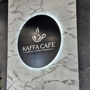 Kaffa Cafe - Sign - A black circular sign featuring a stylized white coffee cup with steam rising from it, along with the text 'KAFFA CAFE' and the tagline 'where coffee began,' is mounted on a marble wall with a backlit glow. The sign indicates the location of Kaffa Cafe, a coffee establishment. - Cafes Near Me - Crown Point, Indiana