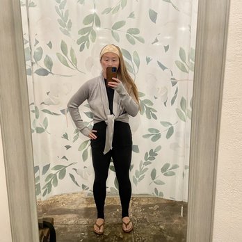 Under the Willow - Gray Ballerina Wrap from Lovestitch in Size Small - A woman is taking a mirror selfie in a fitting room with a leafy-patterned curtain as the background. She has long hair, wears a beige headband, a gray cardigan tied at the waist, black leggings, and flip-flops. The fitting room has a wooden floor, exposed ceiling pipes, and a metal ventilation duct visible at the top. The mirror is framed with a light-colored border, and a small bag is partially visible at the bottom left corner. - Women's Clothing Near Me - Temecula, California