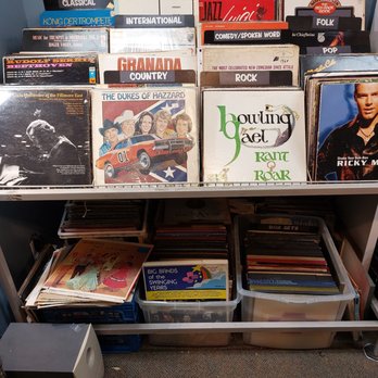 The Second Show - Variety of records - The image shows a display of vinyl records arranged on metal racks and in plastic storage bins. The records are organized by genre, with labels such as Classical, Operatic, Jazz, Folk, and Rock. Some album covers are visible, including a black-and-white photo of a musician, a cartoon illustration of The Dukes of Hazzard, and a portrait of a male artist. The setup appears to be in a record shop or a dedicated music section, with a carpeted floor and a small speaker in the lower left corner. - Thrift Stores Near Me - Hudson, New York