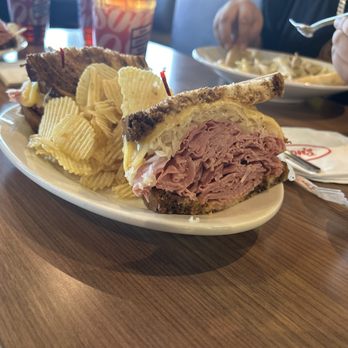 Jason's Deli - Reuben The Great - A close-up of a Reuben sandwich featuring sliced corned beef, melted cheese, and sauerkraut between slices of rye bread, with toasted edges. The sandwich is placed on a white oval plate alongside ridged potato chips. In the background, a person is eating from a bowl, and other food items and drinks are visible on the table, indicating a casual dining setting. - Delis Near Me - Albuquerque, New Mexico