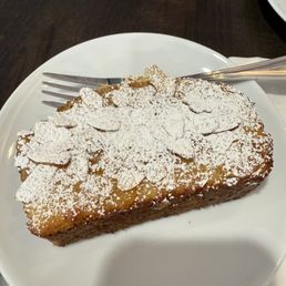 A slice of cake, dusted with powdered sugar, is presented on a white round plate. The cake appears moist and dense, with a golden-brown crust. A metal fork rests on the plate, and part of another plate is visible in the background. The setting suggests a dining environment, possibly a café or restaurant.