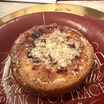 Baker 305 - Egg quiche - A baked egg quiche with a golden-brown crust and a slightly uneven surface, topped with grated cheese. The quiche is placed on a decorative plate featuring religious text and a gold-colored border, with a blurred background including a piece of paper and some greenery. - Bakeries Near Me - Miami, Florida