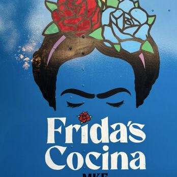 Frida's Cocina - Food Truck - A mural depicts a woman's face with closed eyes, dark eyebrows, and stylized hair adorned with large, colorful flowers in red, yellow, and blue. The background is a solid blue color. Below the face, the text "Frida's Cocina" is prominently displayed in white, with a small red flower graphic above the letter "i". The word "MKE" appears underneath in black. The mural is painted on a flat surface, possibly a vehicle or wall, with visible rivets and a glossy finish. - Tacos Near Me - Milwaukee, Wisconsin