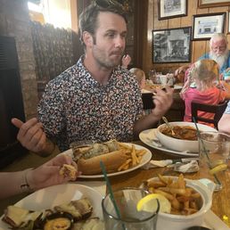 My man, Dan, ordered the French Dip and Guinness Beef Stew! - A man with dark hair, wearing sunglasses on his head and a floral shirt, is seated at a table with various dishes, including French fries, a sandwich, and a bowl of stew. The restaurant has wood-paneled walls decorated with framed pictures, and other diners, including children and an older man with a beard, are visible in the background.