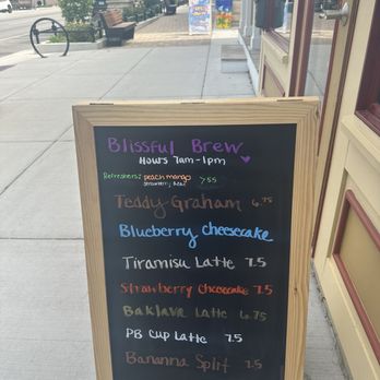 Blissful Brew Coffee Co - A chalkboard sign outside Blissful Brew Coffee Co features handwritten menu items including flavored lattes, cheesecakes, and other drinks, with prices listed. The sign is placed on a sidewalk near the storefront, with a street scene in the background showing benches, trees, and storefronts. The menu includes options like Blueberry Cheesecake, Strawberry Cheesecake, and Banana Split, with prices ranging from $1.50 to $7.50. - Coffee & Tea Near Me - Lockport, Illinois