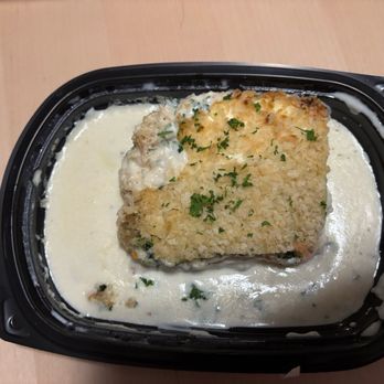 Wegmans - Not a fan of the Veggie Lasagna, bland and soggy veggies - A breaded fish fillet, garnished with chopped herbs, is positioned in a black plastic container filled with a creamy white sauce. The container appears to be for takeout or prepared food, with the fish partially submerged in the sauce. - Grocery Near Me - Tysons, Virginia
