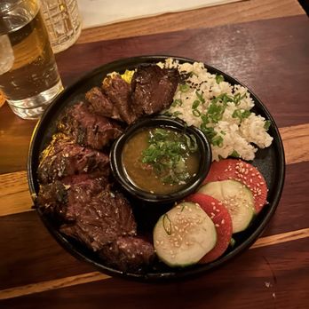Suzume - Steak plate - A steak plate served with rice, sliced cucumbers and tomatoes, and a dipping sauce, on a dark round plate at a restaurant. - Ramen Near Me - Brooklyn, New York