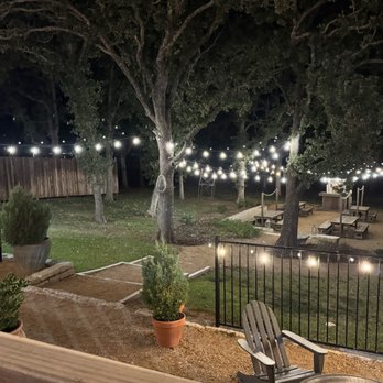 At The Table - Outdoor area - Outdoor area illuminated by string lights hanging between trees, with picnic tables and benches arranged for social gatherings. Potted plants and a wooden fence are visible, creating a designated space for events or casual outdoor activities. The scene is captured at night, emphasizing the lighting and natural setting. - Community Service/Non-Profit Near Me - Argyle, Texas