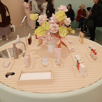 Glossier Chicago - A round display table in a beauty store features skincare products, a floral arrangement, and informational labels, with customers in the background. - Cosmetics & Beauty Supply Near Me - Chicago, Illinois