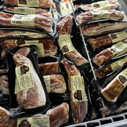 Bacon - Various packages of smoked bacon are arranged in a refrigerated display, each with labels indicating the product type and weight.