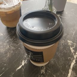 Decaf latte - A disposable coffee cup with a black plastic lid and a cardboard sleeve is positioned on a dark marble table. In the background, there is a clear plastic cup containing iced coffee, and a small glass vase with a green plant. The setting appears to be inside a cafe or coffee shop, with natural light coming through large windows.