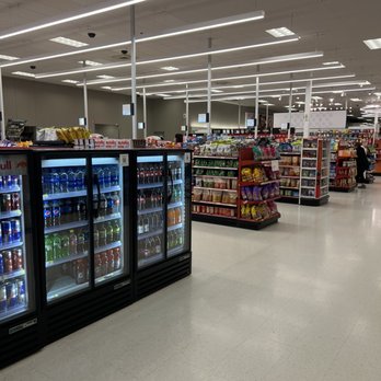 Target - The image shows an interior of a grocery store aisle with refrigerated drink coolers containing bottled beverages, including sodas and water. Shelves with snack products such as chips are visible further back. The ceiling has bright, evenly spaced linear LED lights, and digital screens are mounted above the aisle. The floor is light-colored tile, and a few shoppers are seen in the distance. - Department Stores Near Me - Hackensack, New Jersey