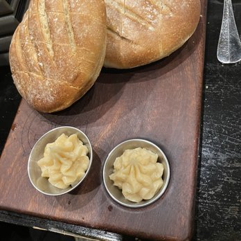 ChopHouse Grille - Complimentary bread along with cinnamon butter. - Two oval-shaped loaves of bread with a golden-brown crust and scoring on top are arranged on a rectangular wooden serving board. Each loaf has a dusting of flour on the surface. Two small metal cups contain a creamy, pale yellow butter, positioned in front of the bread. The setup is on a dark table, with a metal utensil and a napkin visible nearby. - New American Near Me - Wakefield, Rhode Island
