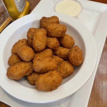 Irish Times - Cheese Curds - A white plate contains multiple fried cheese curds, which are golden-brown and crispy. The plate is on a white tray, with a small cup of creamy dipping sauce nearby. The background includes a wooden table and part of a yellow condiment bottle. - Pubs Near Me - Brookfield, Illinois
