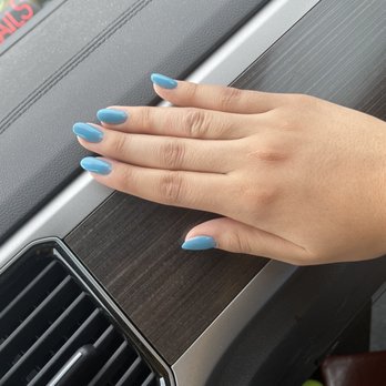Red Nails - All on natural nail except for the pointer finger which they added $5 just for a short fake nail. Bad nail shape and crooked - A hand with five fingers, featuring blue painted nails, is resting on a car dashboard that has a wood-like finish. The dashboard includes an air vent with black slats and a silver trim. The background shows part of a car window and a blurred exterior scene. The image focuses on the hand and the interior details of the vehicle. - Nail Salons Near Me - Eagan, Minnesota