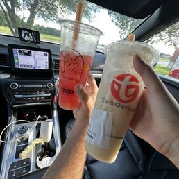 Sea Salt Jasmine Tea - Inside a car, two individuals hold up large plastic cups with beverages, one with a pink drink and the other with a light-colored drink. The car's dashboard, GPS navigation screen, and rearview mirror are visible, along with a small mounted device. The background shows trees and a partly cloudy sky through the windshield, suggesting a daytime drive.