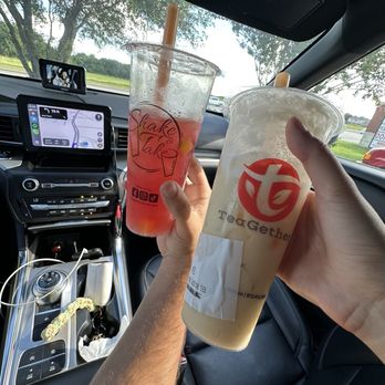 Teagether - Mango dragonfruit lemonade and sea salt jasmine tea - Inside a car, two individuals hold up large plastic cups with beverages, one with a pink drink and the other with a light-colored drink. The car's dashboard, GPS navigation screen, and rearview mirror are visible, along with a small mounted device. The background shows trees and a partly cloudy sky through the windshield, suggesting a daytime drive. - Bubble Tea Near Me - Grand Prairie, Texas