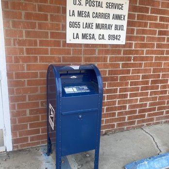 US Post Office - Ok - A blue USPS mailbox stands on the sidewalk in front of a brick wall. Above it, a white sign displays the address for the U.S. Postal Service, La Mesa Carrier Annex, located at 6055 Lake Murray Blvd, La Mesa, CA 91942. The mailbox is positioned near a curb with a blue painted parking space line and a wheel stop block nearby. - Post Offices Near Me - La Mesa, California
