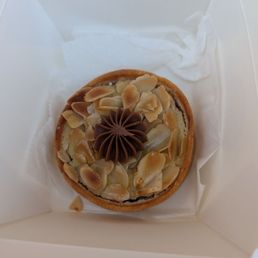 Tarts - A round tart topped with sliced almonds and a central chocolate decoration in a swirl pattern is shown inside a white box. The tart appears to have a pastry crust and is presented on a piece of tissue paper within the box, which is open to display the dessert.