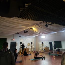 Collective Flow MKE - A yoga class is taking place in a studio with wooden floors, decorative string lights, and fabric draped from the ceiling. Participants are seated on mats, some stretching, with a DJ setup and speakers at the front.