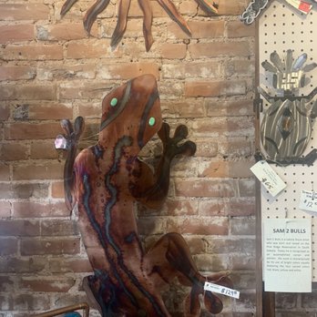 Backroads Art - A large wooden gecko sculpture with glowing green eyes is mounted on a brick wall, with other metal art pieces nearby. - Art Galleries Near Me - Weston, Missouri