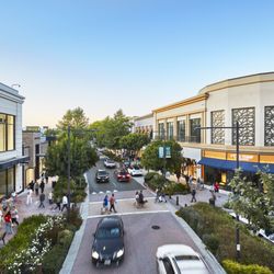Shoppers and cars move along a tree-lined street between Zara and L'Occitane at Broadway Plaza.
