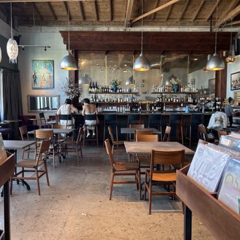 Folklore - Bar - A cafe or bar interior with a vinyl record display in the foreground, wooden tables and chairs, a bar with bottles, and two people sitting at the bar. - Vinyl Records Near Me - Napa, California