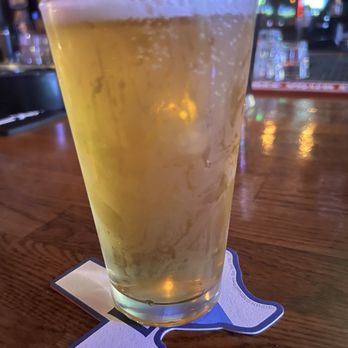 Hideout Pub - A tall glass of beer with condensation sits on a bar counter, with a blurred bar background and colorful lights. - Pubs Near Me - Austin, Texas