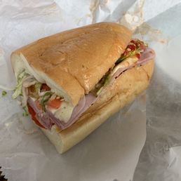 Loaded Regatta with hots - A sandwich with sliced cold cuts, shredded lettuce, tomato slices, and condiments inside a soft, toasted hoagie roll. The sandwich is partially wrapped in white paper, with some ingredients visible from the side. The image focuses on the sandwich, highlighting its fresh ingredients and bread.