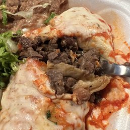 The steak burrito is all meat!! Hope you're hungry - Close-up of cheese-covered enchiladas filled with beef, topped with melted cheese and sauce. The dish is served with side items such as refried beans and a small salad with greens. The food is presented on a foam takeout container, indicating a casual dining or takeout setting.