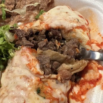 Que Rico - The steak burrito is all meat!! Hope you're hungry - Close-up of cheese-covered enchiladas filled with beef, topped with melted cheese and sauce. The dish is served with side items such as refried beans and a small salad with greens. The food is presented on a foam takeout container, indicating a casual dining or takeout setting. - Mexican Near Me - Chicago, Illinois