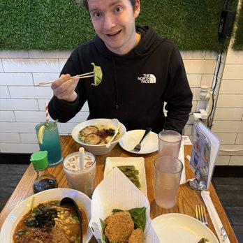 Buntopia - A young man with light brown hair, wearing a black North Face hoodie, is seated at a table with Asian cuisine, including ramen and sushi, in a restaurant setting. - Asian Fusion Near Me - Brooklyn, New York