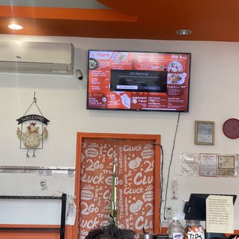 Cluck2Go - Front counter w menu - The image shows the interior of a restaurant with a wall-mounted flat-screen TV displaying a menu. An air conditioning unit is installed above the TV. To the left, a decorative chicken sign with a 'WELCOME' banner hangs on the wall. The wall is painted white, with an orange ceiling accent. Part of a counter with a hand sanitizer bottle and a cash register is visible at the bottom right. Several notices and a red clock are also on the wall. - Singaporean Near Me - Pasadena, California