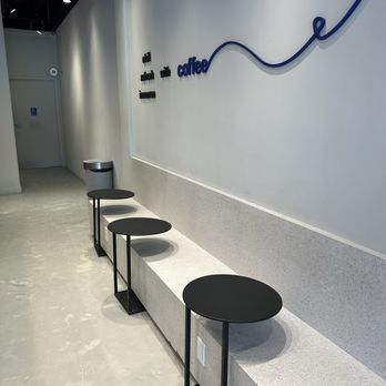 A Lot Coffee - Seating area - A row of small black round tables with black metal legs is positioned along a white wall in a coffee shop, with a trash can nearby. - Coffee & Tea Near Me - Markham, Ontario