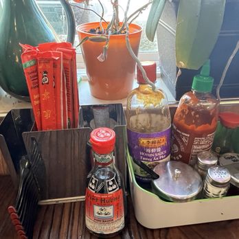 Pho Saigon - Table condiments - The image shows a restaurant table with a variety of condiments, including sauces, spices, and utensils, arranged in a container. There are also chopsticks and a bottle of fish sauce. The background features a window with potted plants and natural light, and an electrical outlet is visible on the wall. - Soup Near Me - Dover, Delaware