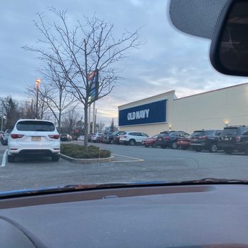 Old Navy - Sign - The photo captures the view from inside a car, looking out through the windshield at an Old Navy store with a large blue sign. The parking lot is filled with various parked vehicles, and a leafless tree is visible near the store. The sky is overcast, indicating a cloudy day, and the scene appears to be in a suburban shopping area. - Men's Clothing Near Me - Clifton, New Jersey
