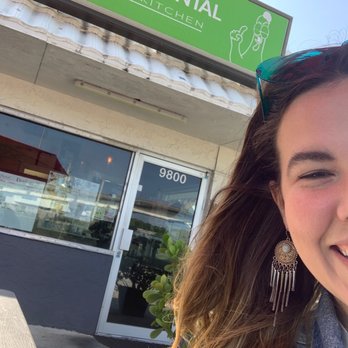 Cafe Oriental - Front entrance of restaurant and a Happy Customer - The image shows a woman smiling with only part of her face visible, wearing earrings and sunglasses on her head. She is outdoors in front of Café Oriental Asian Kitchen, which has a green sign with white text and a line drawing of a woman holding a spoon. The café's entrance has a glass door with the number 9800 above it, and there is a potted plant near the door. A red umbrella is partially visible at the top of the image. - Asian Fusion Near Me - Miami, Florida