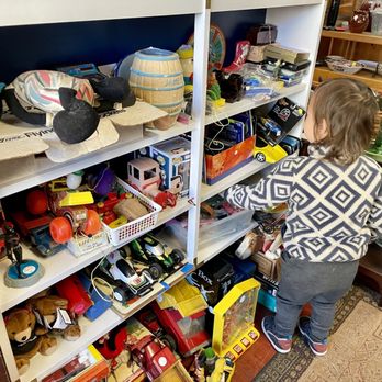 Kingston Consignments - Checking out some toys - A young child with shoulder-length hair, dressed in a patterned sweater and jeans, is standing in front of a white shelving unit filled with various toys, collectibles, and miscellaneous items. The store has wooden floors, and other furniture and decorative items are visible in the background. The child appears to be examining the toys on the shelf. - Antiques Near Me - Kingston, New York