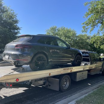 That Guy Towing - Solid strap placement! - A black Porsche Macan is secured on a flatbed tow truck on a sunny residential street with trees and parked cars. - Towing Near Me