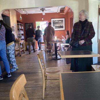 Raging Sage - The image shows the interior of a coffee shop with several customers. A woman with white hair, wearing a plaid jacket and carrying a black purse, stands near a table on the right side. In the background, other customers are near the entrance, some standing and some seated, with one person using a phone. The decor includes a framed black-and-white photograph on a reddish wall, wooden tables and chairs, and a ceiling fan. The space has a warm, inviting atmosphere with natural light coming from the entrance. - Coffee & Tea Near Me - Tucson, Arizona