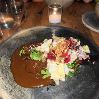 Claro - Pork Cheek Mole Rojo - A dish featuring a meat portion with brown sauce, served alongside a mixed salad with fried onions, herbs, and radicchio on a black plate. - Mexican Near Me - Brooklyn, New York