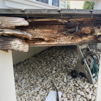 Sunburst Construction - Not pressure treated wood - A wooden deck with severely rotted and broken wood planks, exposing the underlying structure. The wood appears weathered and deteriorated, indicating potential structural issues. The deck is elevated above a gravel ground, with a garden hose reel visible underneath. The background shows part of a house with siding and windows. - General Contractors Near Me - Leesburg, Virginia